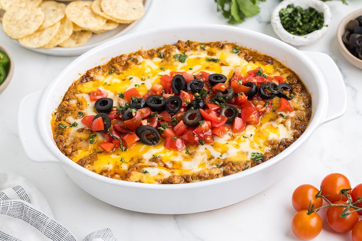 Cheesy taco dip in a white oval dish topped with black olives and diced tomatoes.