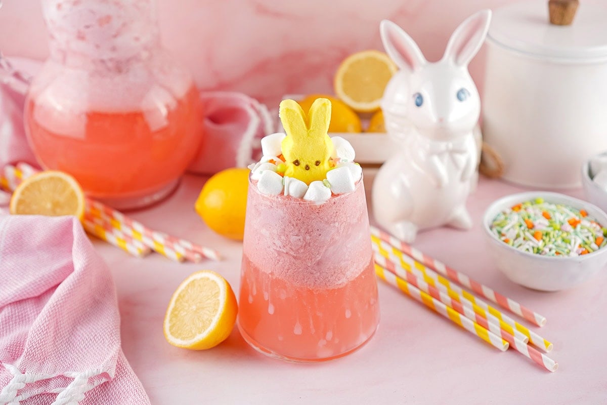 Tall glass of pink lemonade slush, Peeps bunny on top, surrounded by lemons and pink straws.