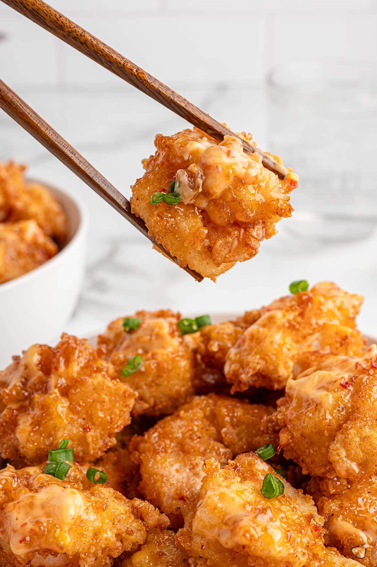 Bowl of golden Bang Bang shrimp with creamy sauce and green onions, chopsticks lift one piece.