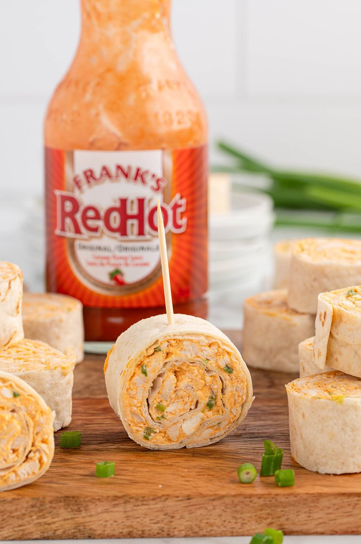 Buffalo chicken pinwheels on a wood board with Frank's RedHot sauce bottle and green onions.
