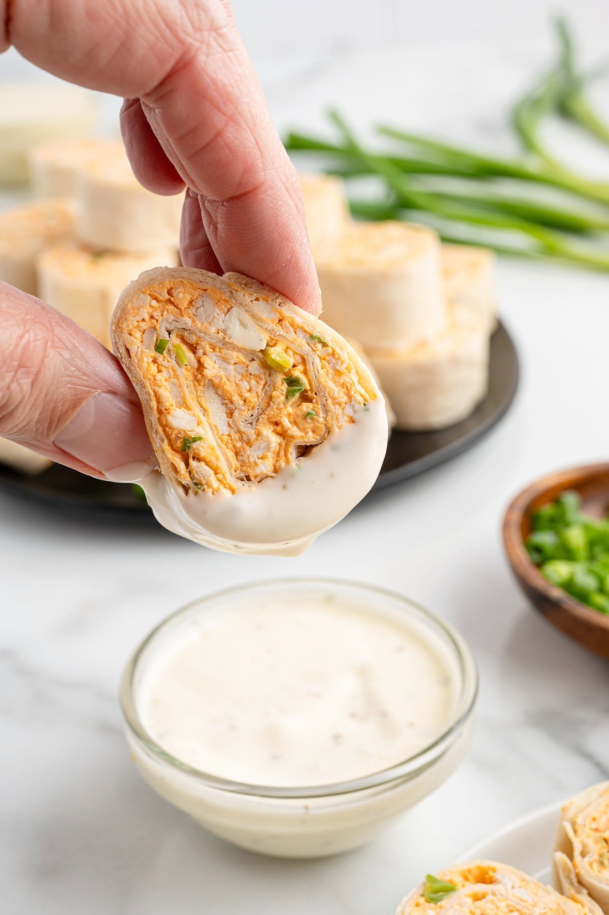 Hand holds a buffalo chicken pinwheel dipped in white sauce, with more on a black plate and green onions nearby.