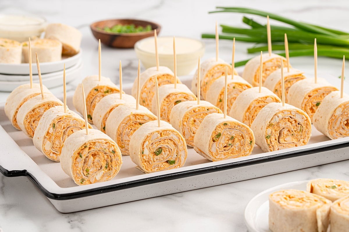Tray of buffalo chicken pinwheels on skewers next to ranch dip and green onions.