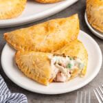 Two golden chicken pot pie hand pies sit on a white plate. One splits open to reveal creamy chicken and peas.
