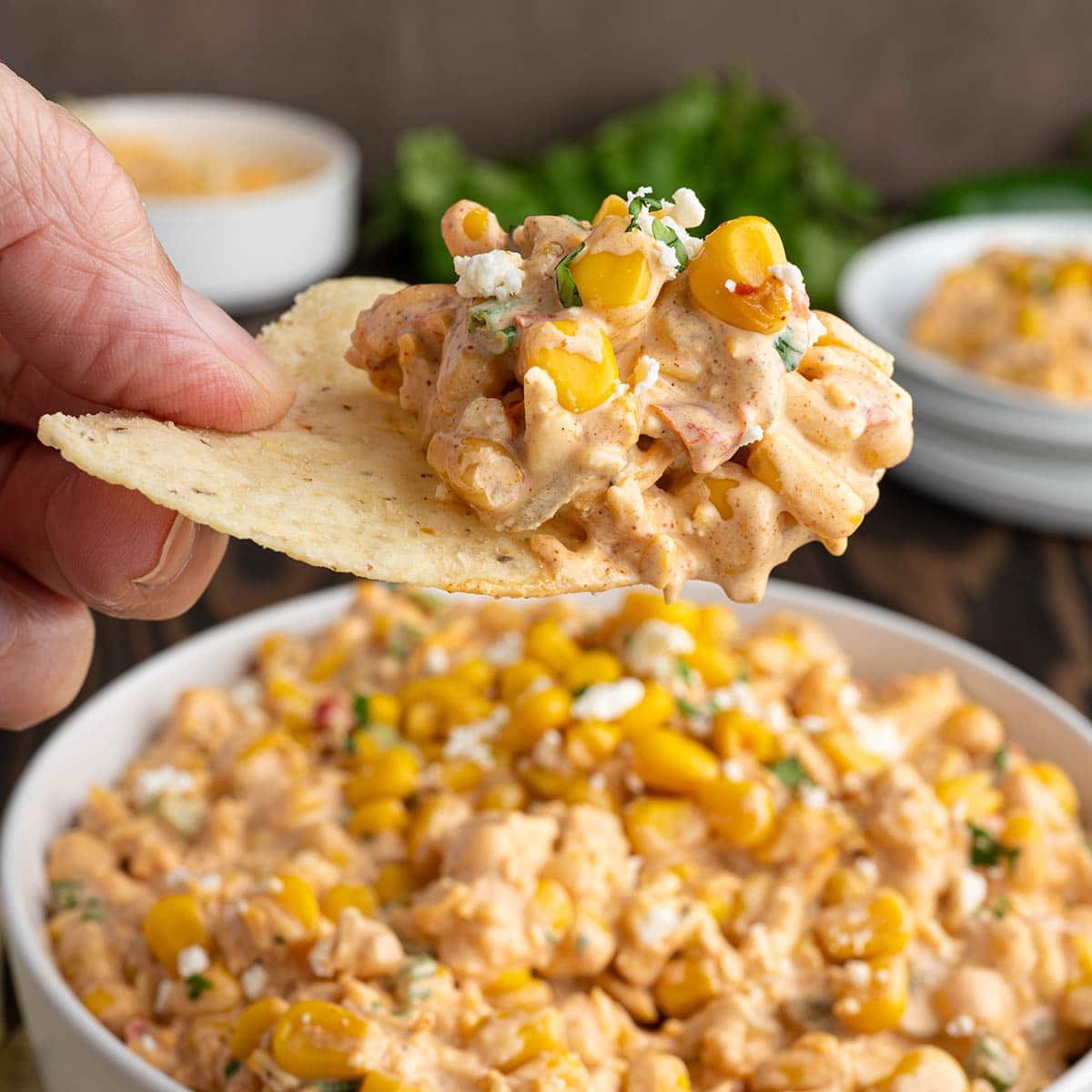 Hand dips tortilla chip into creamy Mexican street corn dip with corn kernels and cilantro.