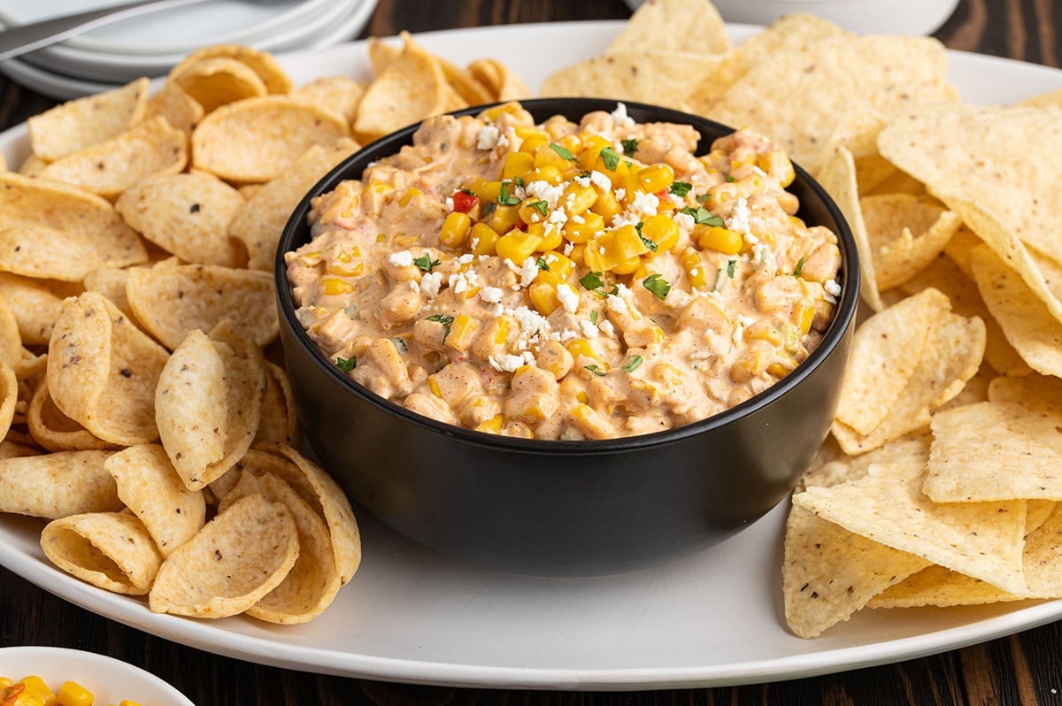 Bowl of Mexican street corn dip with corn kernels, cheese, and cilantro, surrounded by tortilla chips.