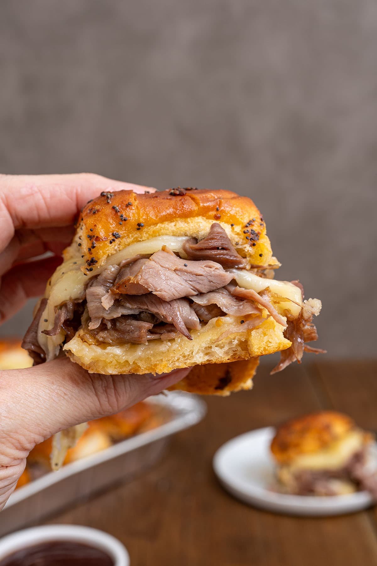 Hand holds a roast beef slider with melted cheese on a sesame seed bun.