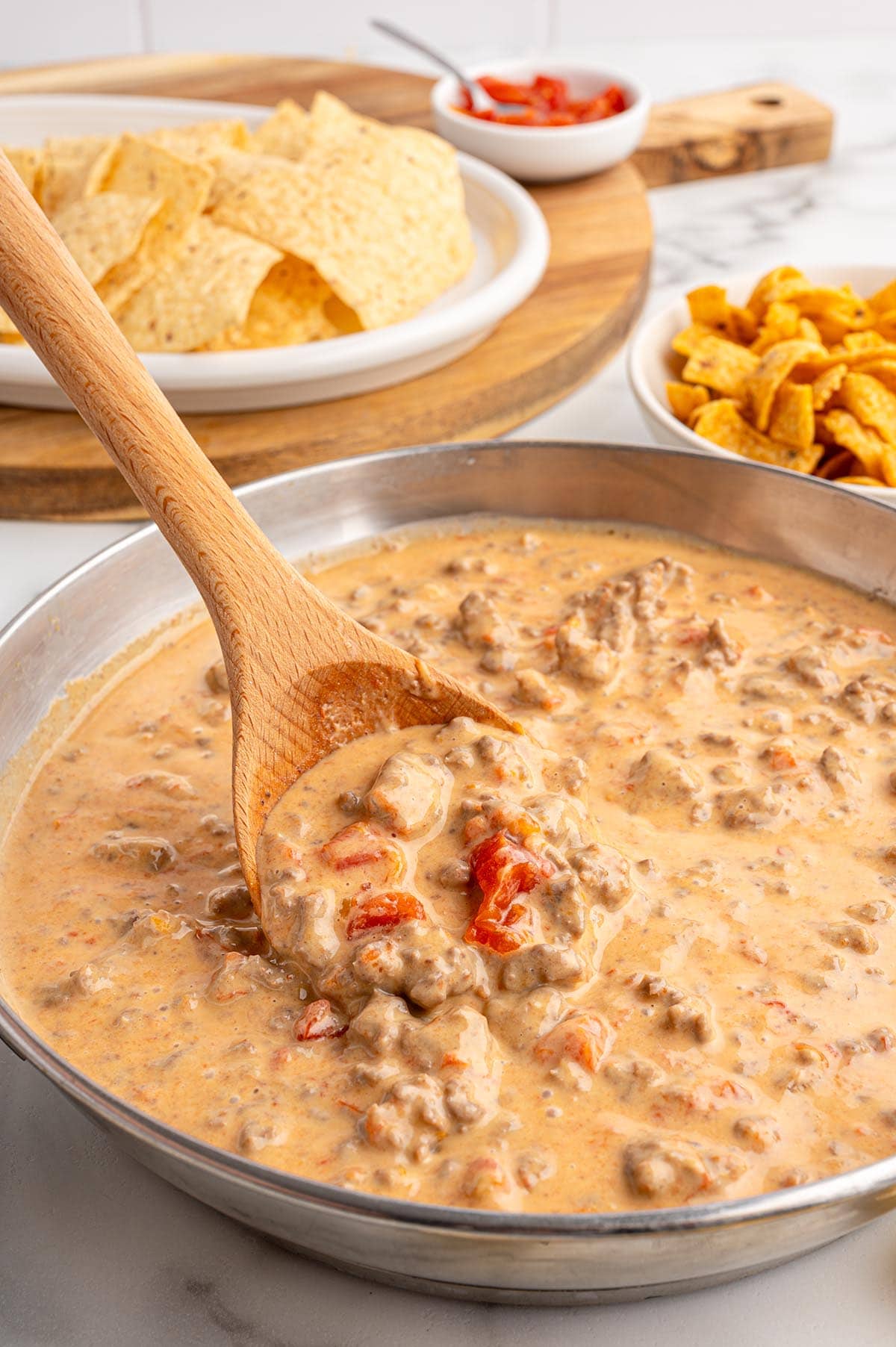 Wooden spoon scoops creamy Rotel dip with ground beef and tomatoes from a silver pot.