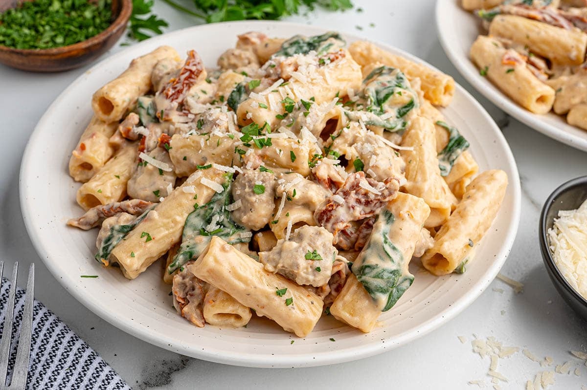 Creamy Italian sausage pasta on a white plate with spinach and sun-dried tomatoes.