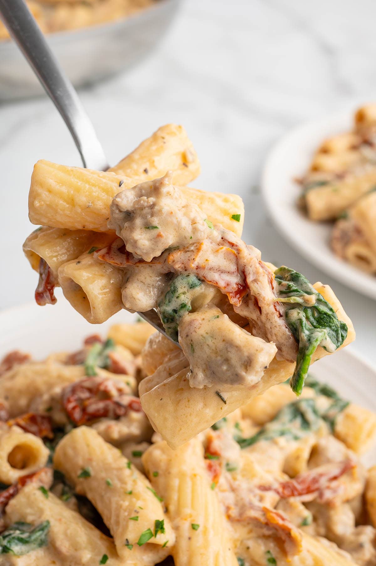 Creamy Italian sausage pasta with rigatoni, spinach, and sun-dried tomatoes on a spoon.