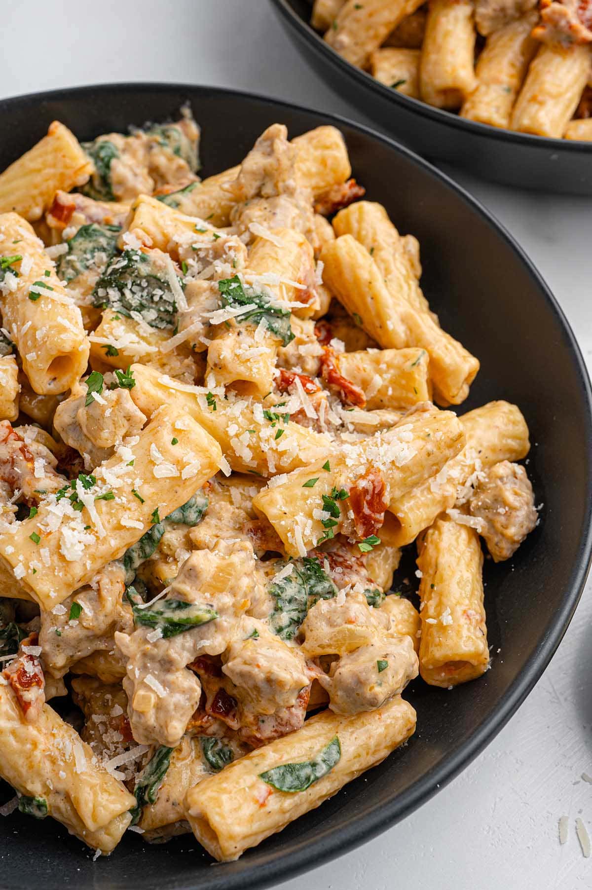 Creamy Italian sausage pasta in a black bowl with spinach, tomatoes, and cheese topping.
