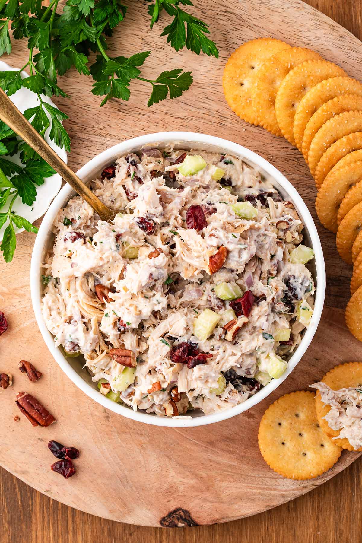 Bowl of cranberry chicken salad with apples, pecans, and parsley garnish next to crackers on a wooden board.