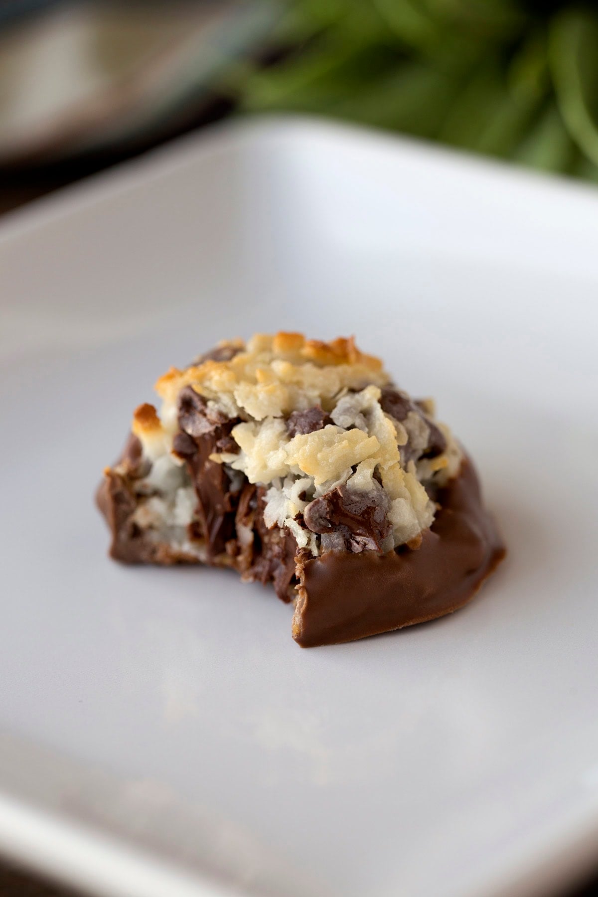 A chocolate chip macaroon sits on a white plate with a fresh bite taken out, revealing coconut and chocolate inside.