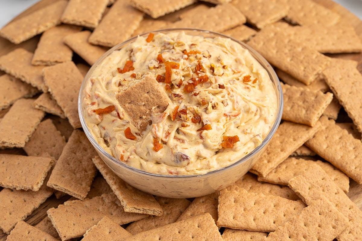 Bowl of creamy carrot cake dip with carrot shreds and nuts, topped with cracker pieces amid stacked wheat crackers.