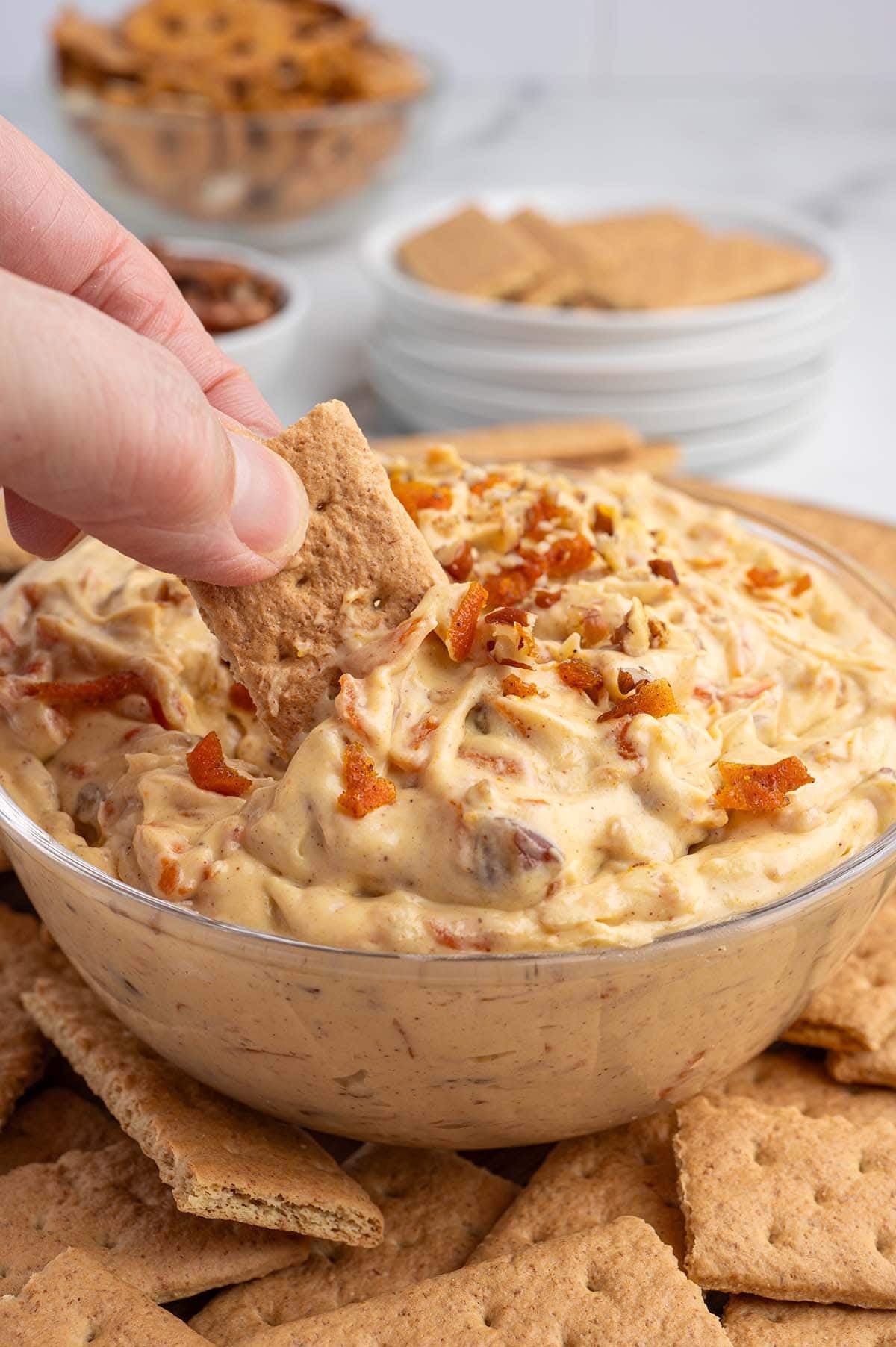 A hand dips a cracker into creamy carrot cake dip with shredded carrots and pecans.