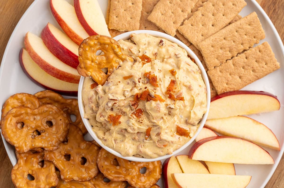 Bowl of carrot cake dip topped with bacon bits, next to apple slices, pretzels, and crackers on a white plate.
