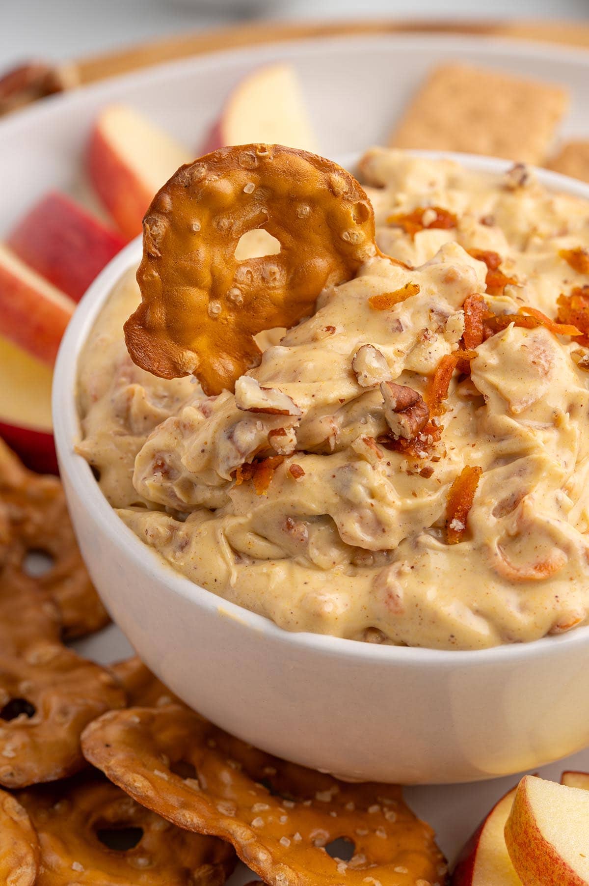 Bowl of creamy carrot cake dip with carrot shreds and pecans, next to apple slices and pretzels.