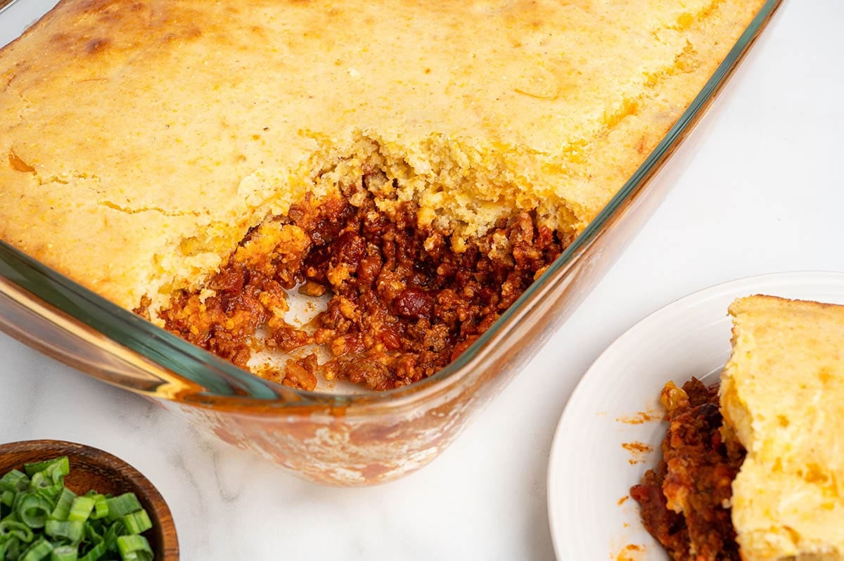 Golden cornbread tops a hearty chili cornbread casserole in a glass dish. A slice sits on a white plate nearby.