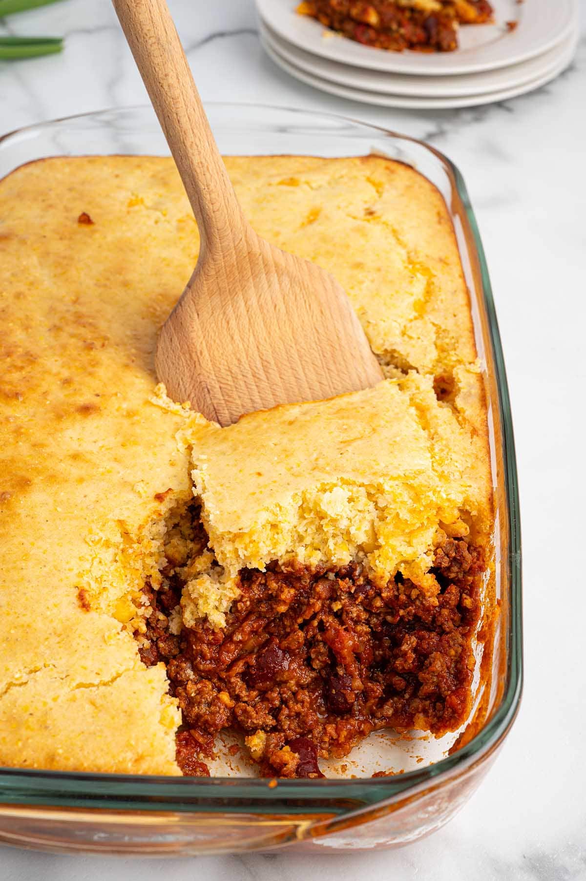 Golden cornbread scoops from a glass dish of chili cornbread casserole. Red chili peeks out below, on a marble counter with green onions.