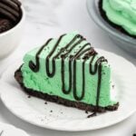 Slice of Grasshopper pie with mint green filling, chocolate drizzle, and Oreo crust on a white plate.