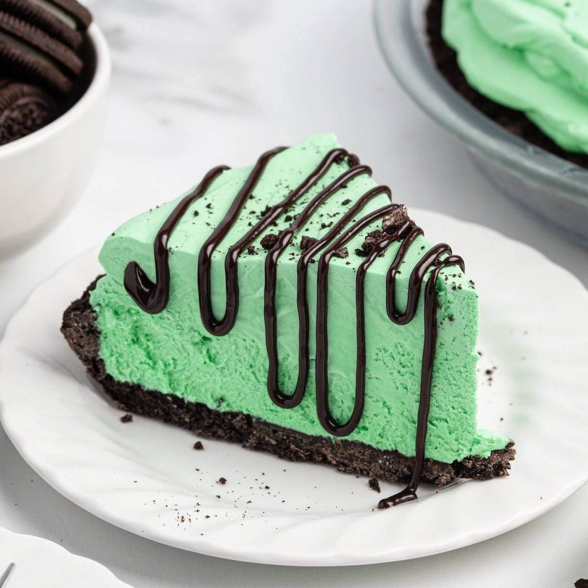 Slice of Grasshopper pie with mint green filling, chocolate drizzle, and Oreo crust on a white plate.