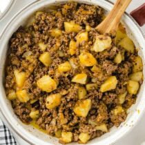 Pot of hamburger hash with ground beef, diced potatoes, and onions in a creamy sauce, wooden spoon inside.