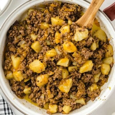 Pot of hamburger hash with ground beef, diced potatoes, and onions in a creamy sauce, wooden spoon inside.