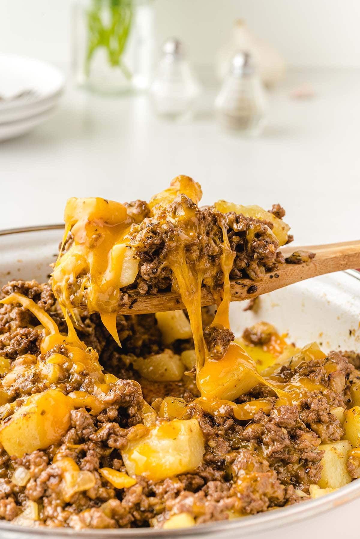 Wooden spoon scoops hamburger hash from pot; melted cheese stretches between spoon and beef-potato mix.