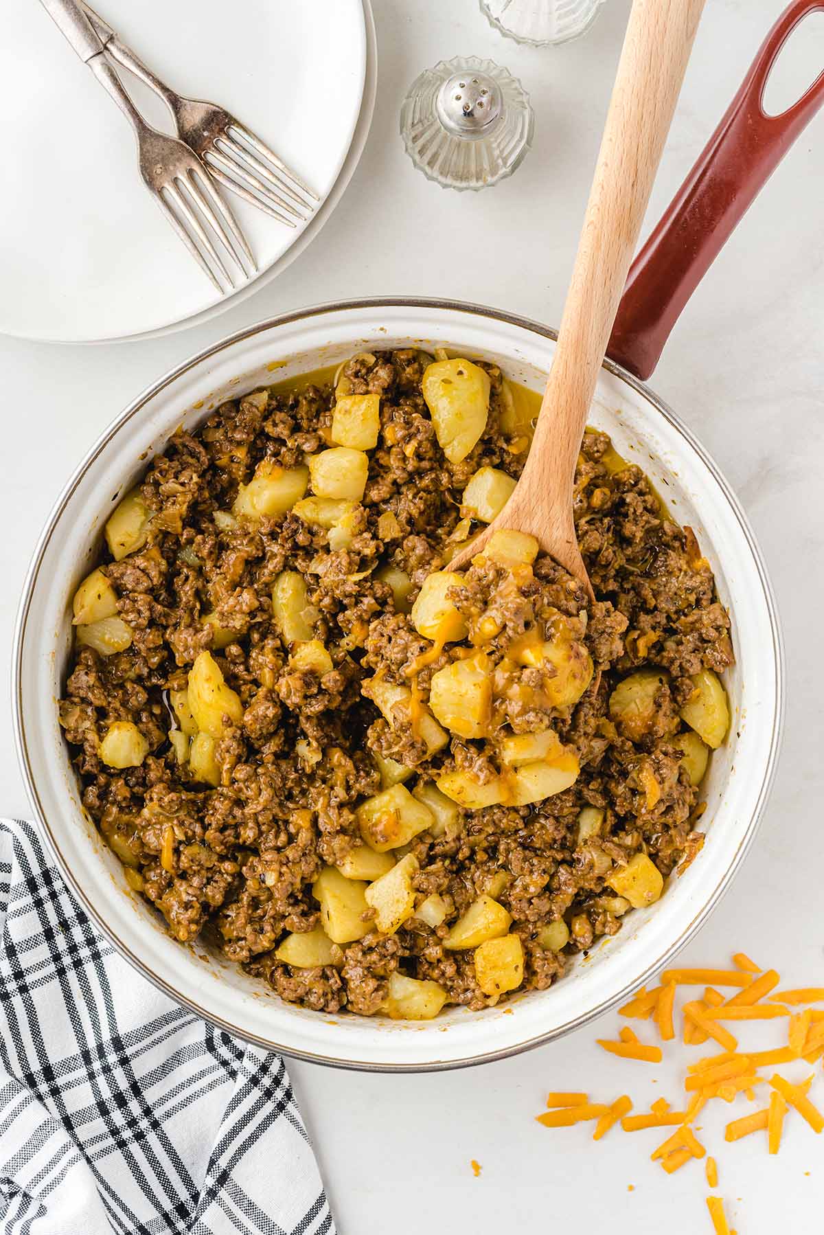 Pan of hamburger hash with ground beef, diced potatoes, and melted cheese; wooden spoon scoops a bite.