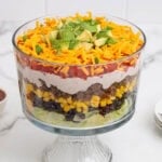 Layered taco salad in a clear glass bowl with lettuce, black beans, corn, ground beef, sour cream, tomatoes, cheese, and avocado.