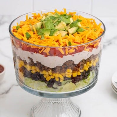 Layered taco salad in a clear glass bowl with lettuce, black beans, corn, ground beef, sour cream, tomatoes, cheese, and avocado.