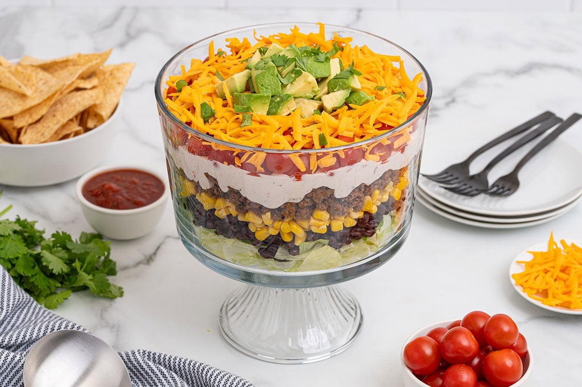 Layered taco salad in a tall glass dish with cheese, avocado, and fresh toppings.
