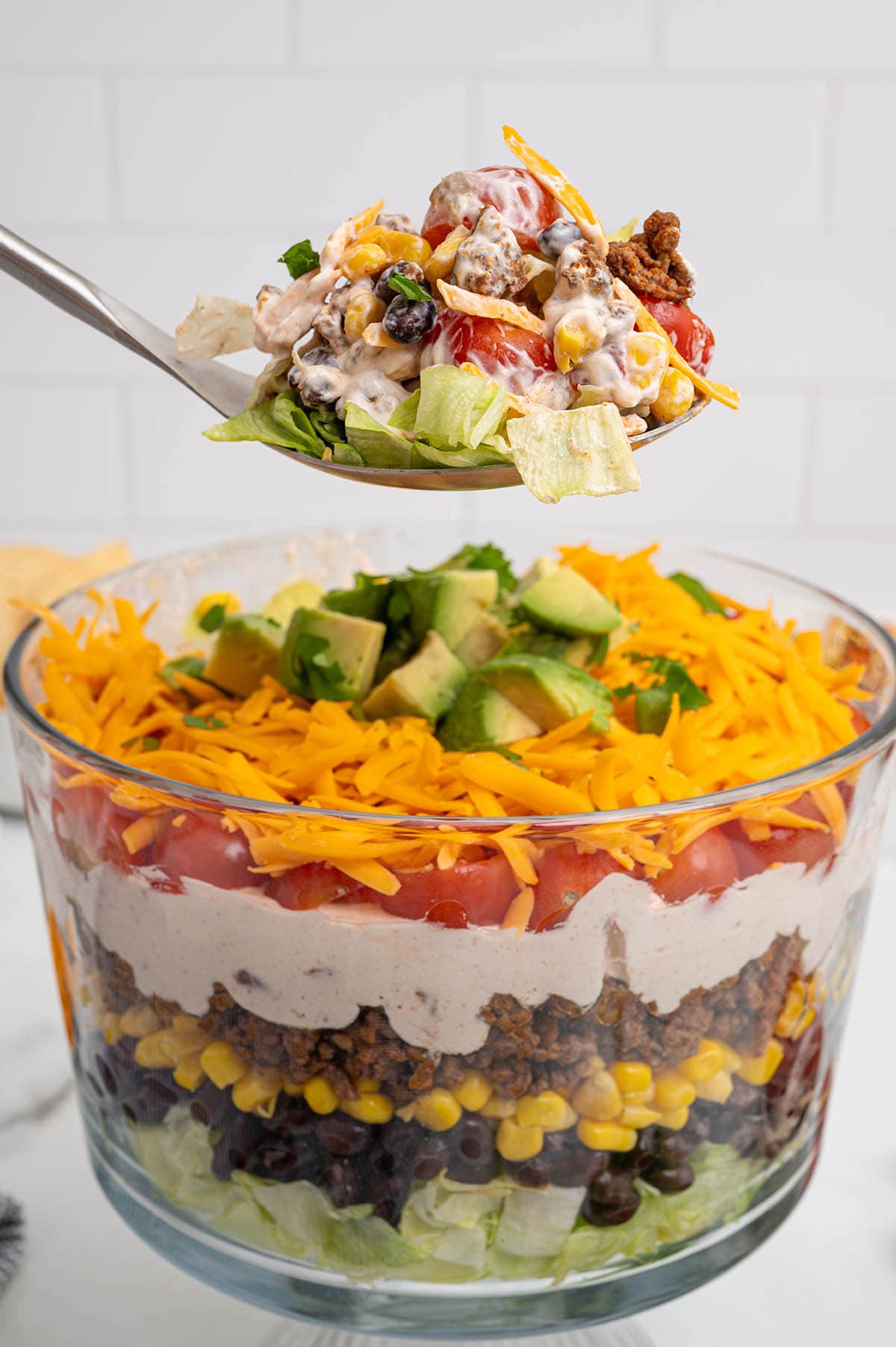 Layered taco salad in a clear glass bowl shows fresh lettuce, beans, corn, beef, sour cream, tomatoes, cheese, and avocado slices on top.