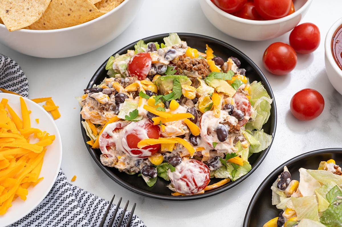 Black bowl holds layered taco salad packed with beef, beans, corn, fresh tomatoes, and melted cheese.