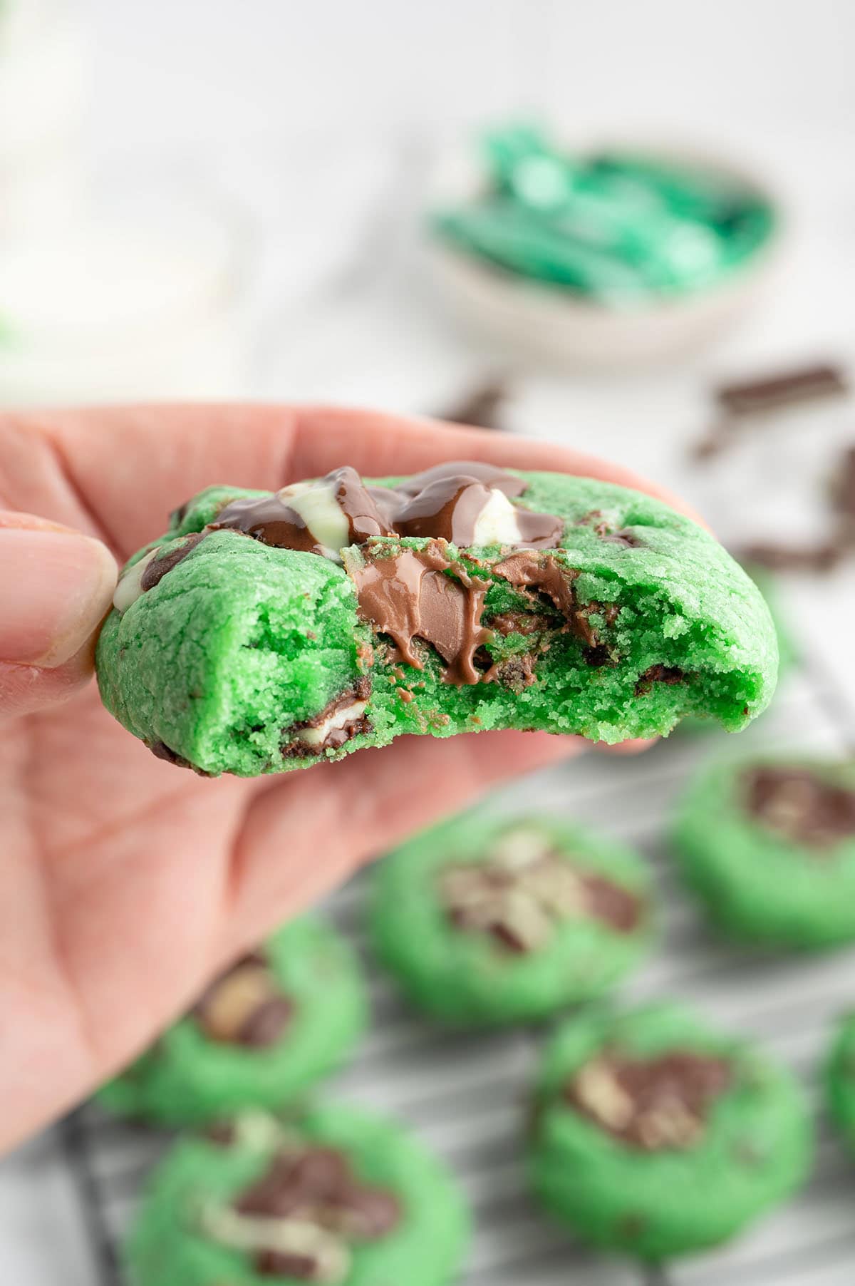Hand holds a bitten green mint chocolate chip cookie with chocolate chunks and white drizzle.