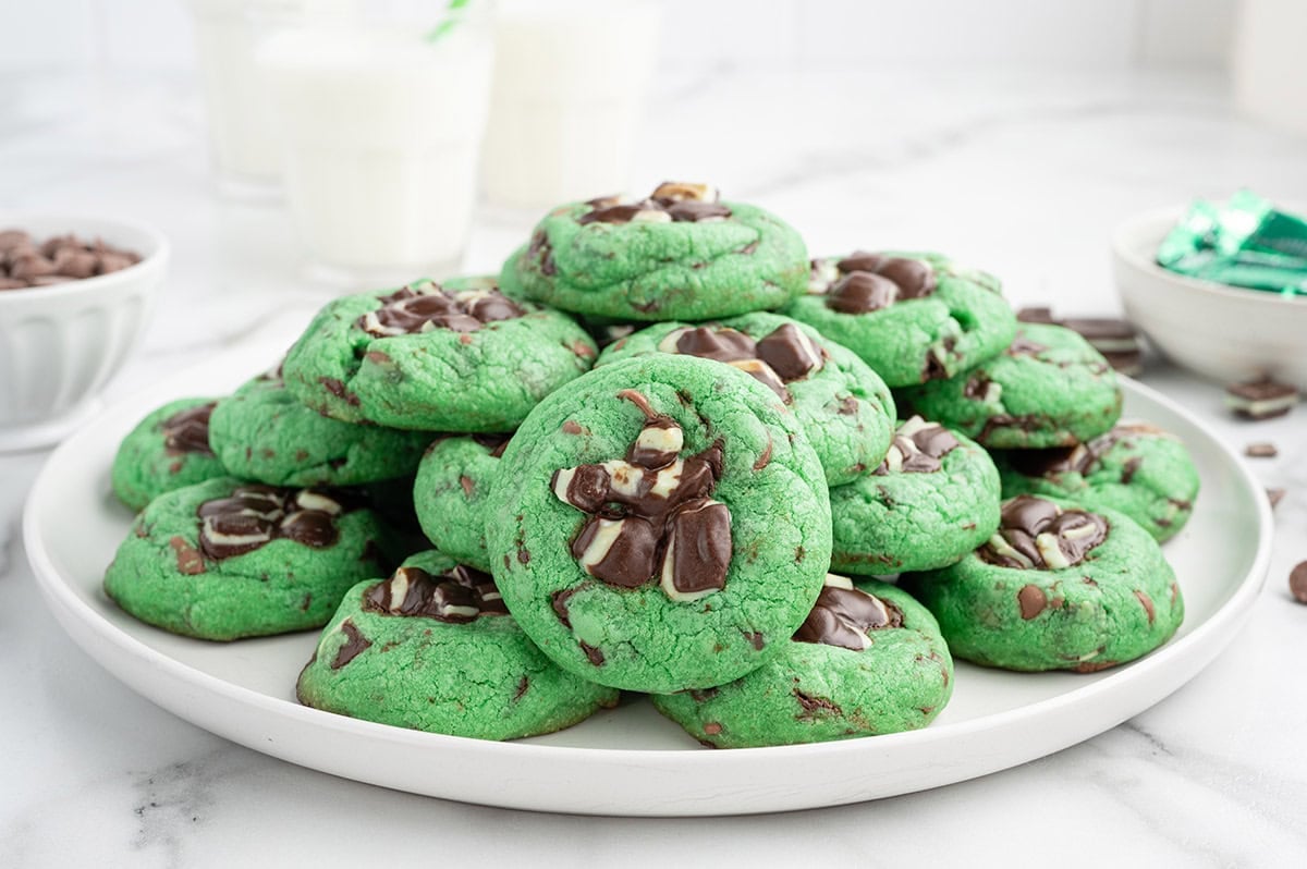 Stack of green mint chocolate chip cookies on a white plate with milk glasses nearby.