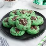 Stack of green mint chocolate chip cookies on a black plate with white chocolate chips and mints in bowls.