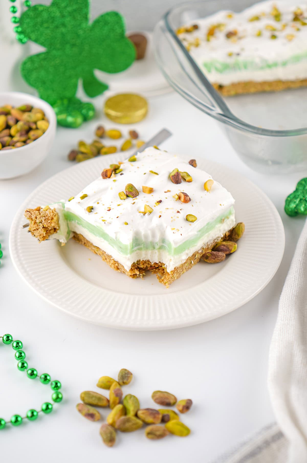 Slice of pistachio pudding dessert on a plate with a bite taken out, topped with whipped cream and pistachios.