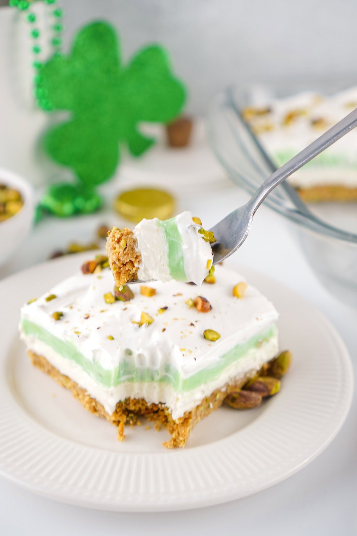 Slice of green pistachio pudding dessert on a white plate, topped with whipped cream and nuts, fork in a bite.