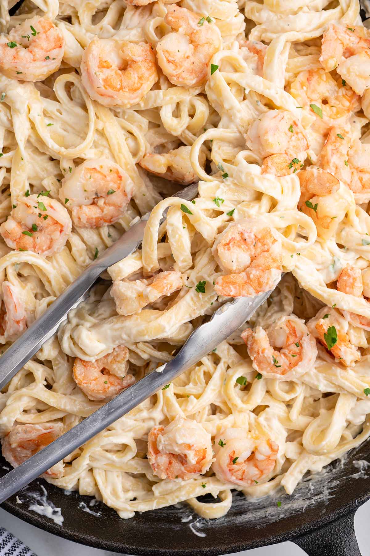 Creamy shrimp alfredo pasta in a cast iron skillet, with tongs lifting noodles and shrimp.