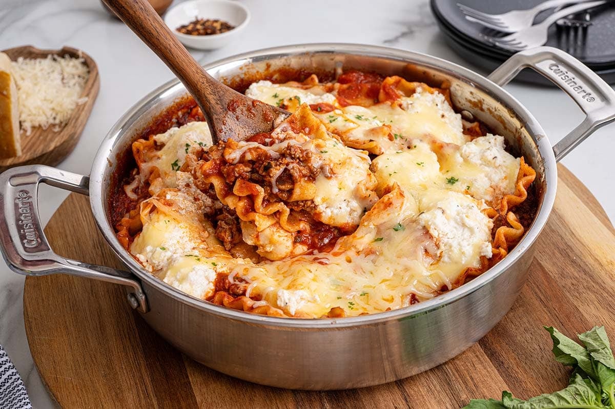 A wooden spoon digging into creamy skillet lasagna.