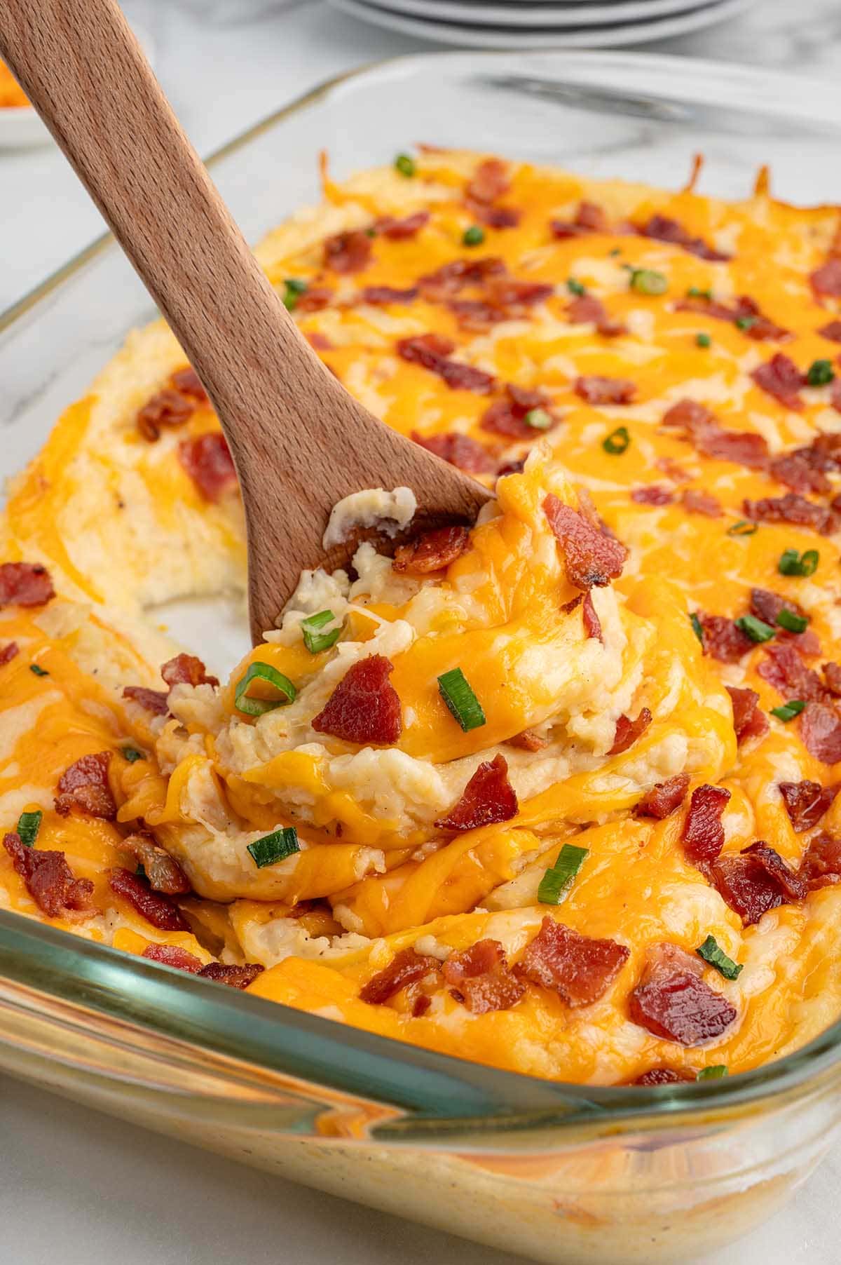 Wooden spoon scoops cheesy twice baked potato casserole loaded with bacon and green onions.