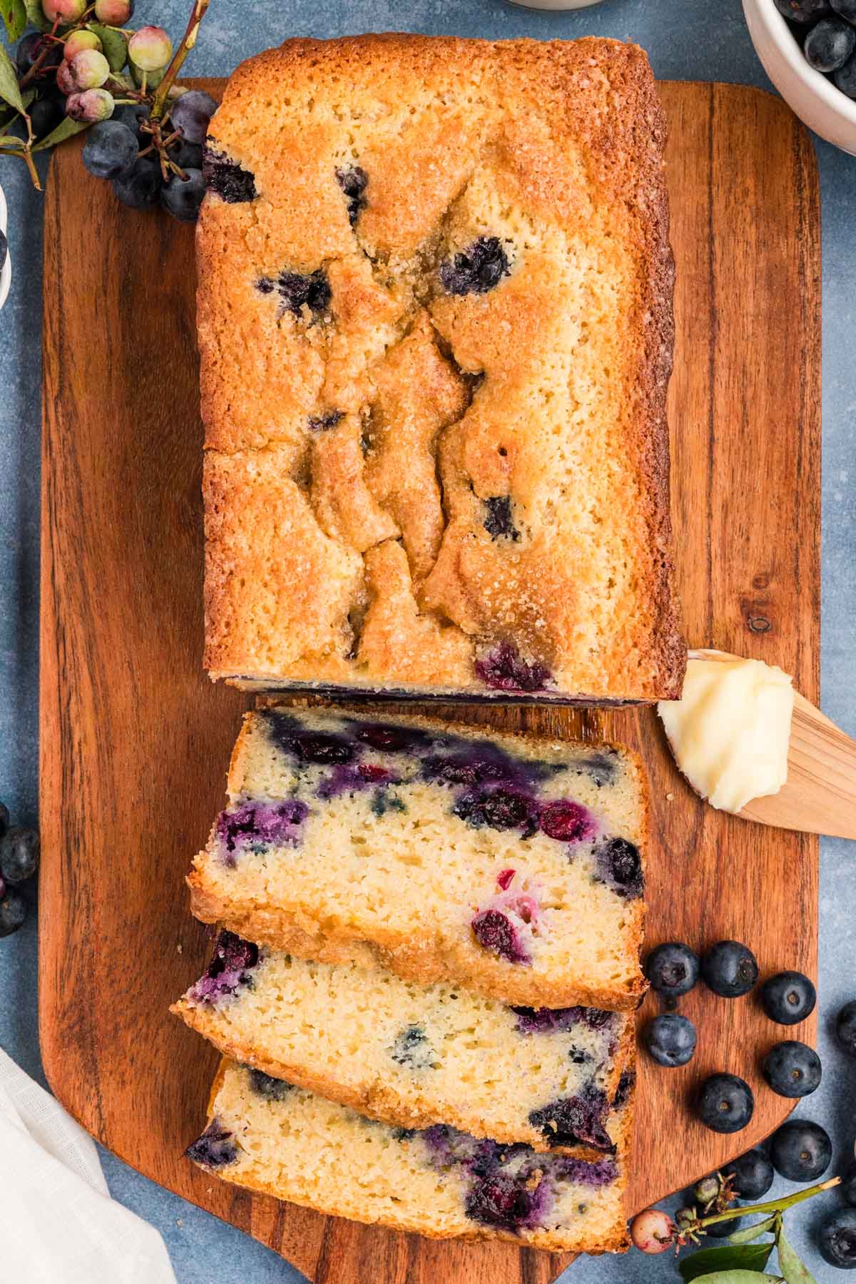 Top view of a blueberry muffin bread loaf with slices cut, showing a golden crust and juicy blueberries throughout on a wooden board.