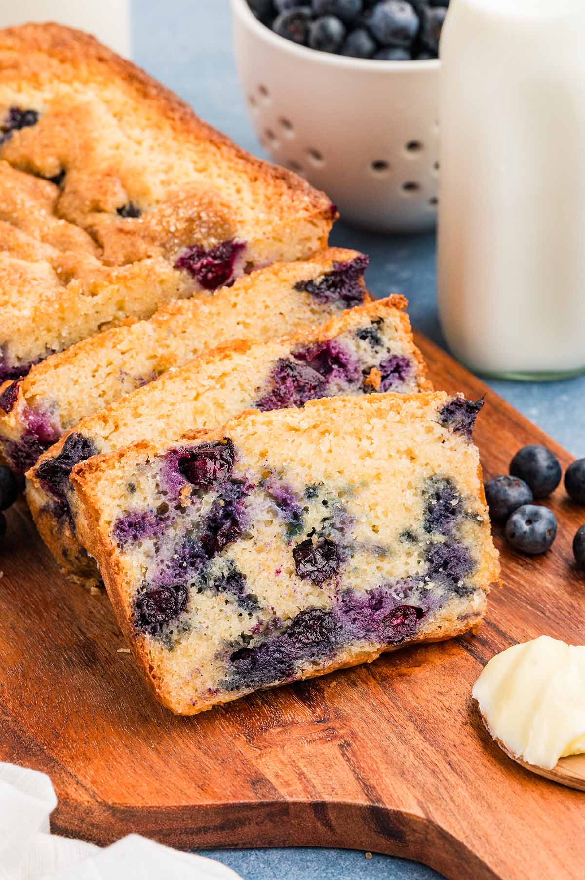 Sliced blueberry muffin bread loaf on a wooden board, showing a moist crumb filled with fresh blueberries, served with milk and extra berries.
