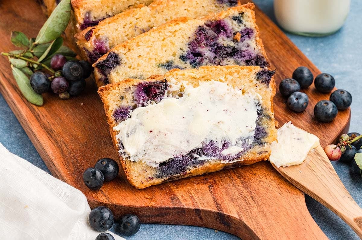 Slices of blueberry muffin bread spread with butter on a wooden board, surrounded by fresh blueberries and a glass of milk.