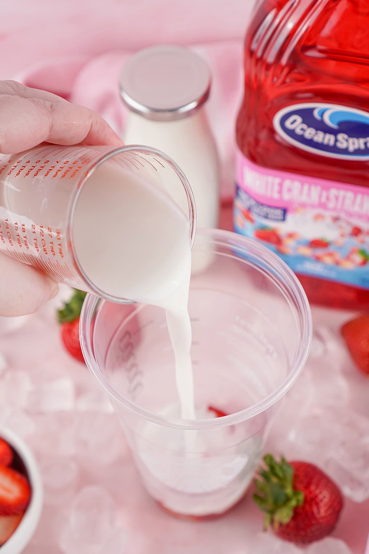 Pouring  coconut milk from a measuring cup into pink strawberry-cranberry juice and fresh berries.