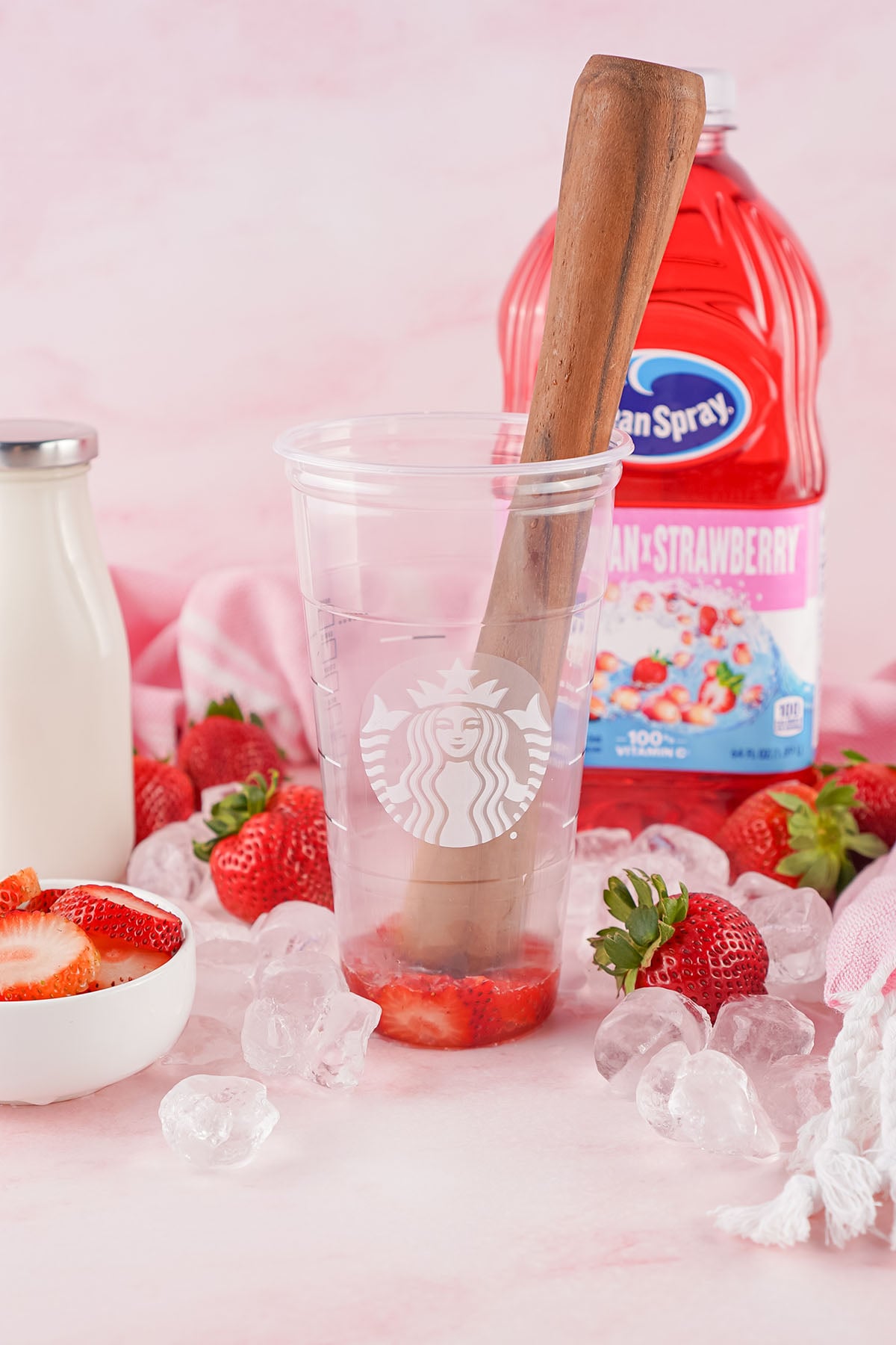 Ingredients for Starbucks Pink Drink: sliced strawberries, ice cubes, pink juice bottle, milk, and muddler arranged on soft pink surface.