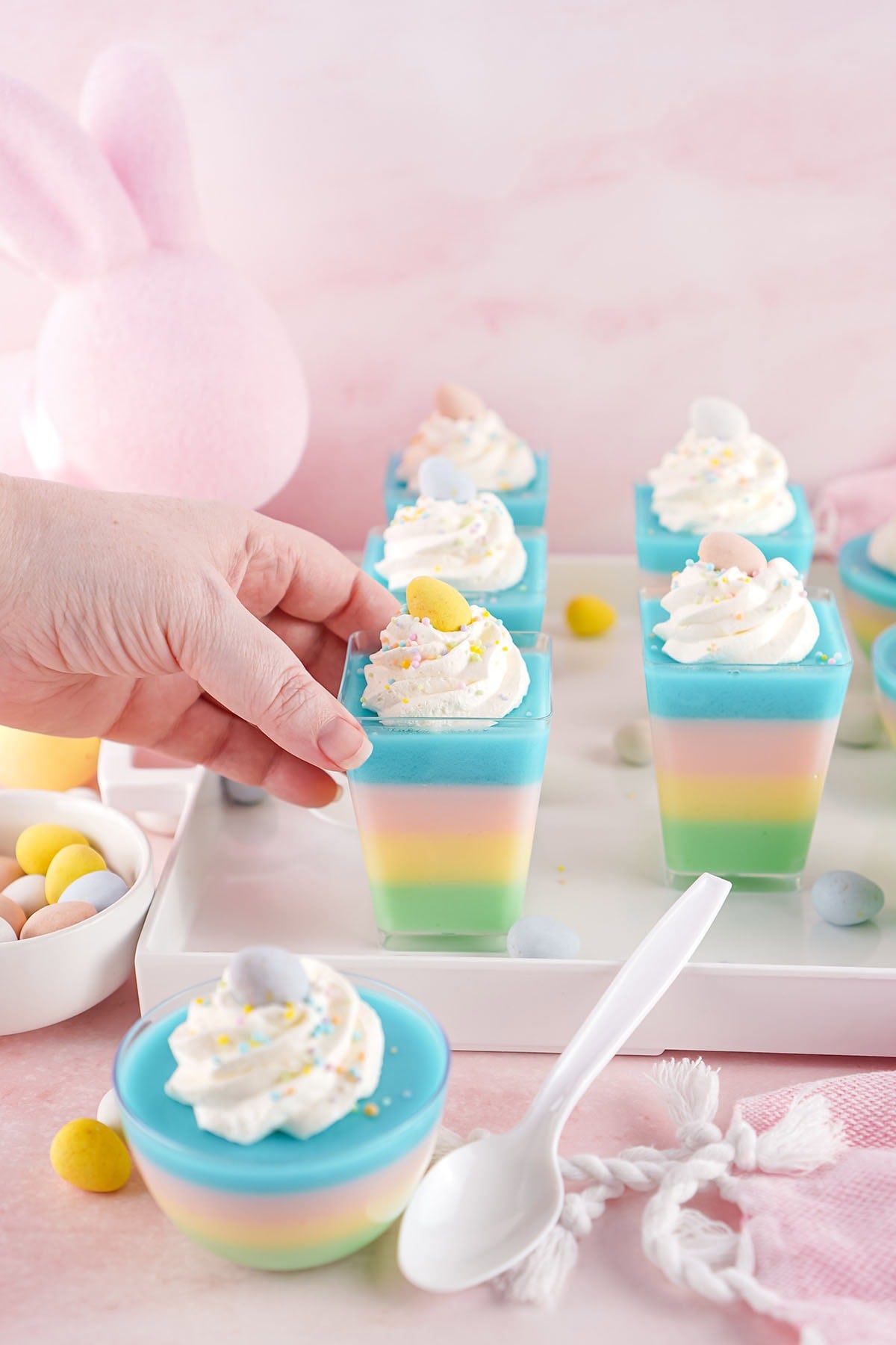 A hand reaching for a clear square cup filled with layered pastel jello, topped with whipped cream and a yellow candy egg, with a pink bunny ear visible in the background.