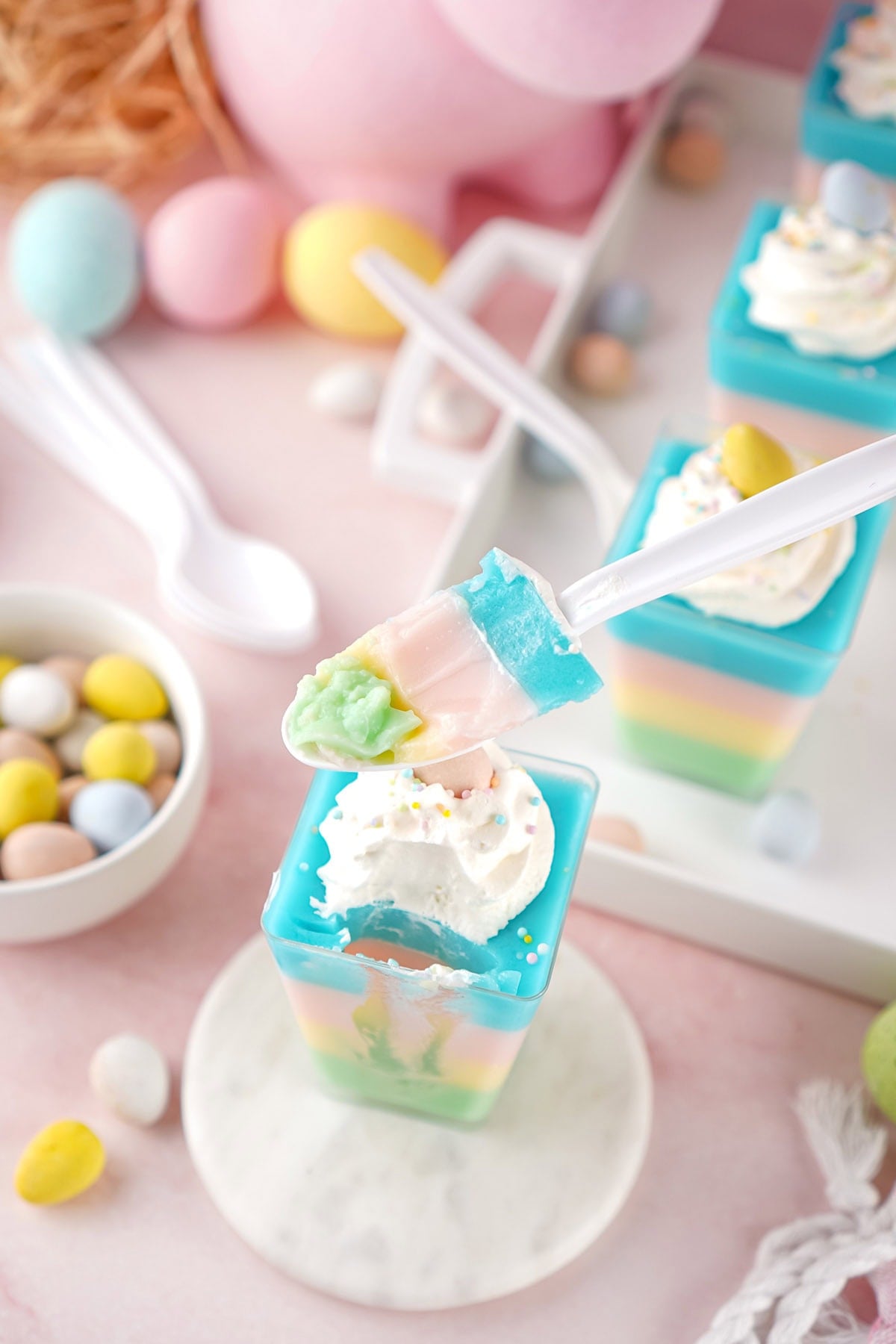 A vertical shot of a white plastic spoon lifting a multi-colored bite of jello, showing the distinct green, yellow, pink, and blue layers.