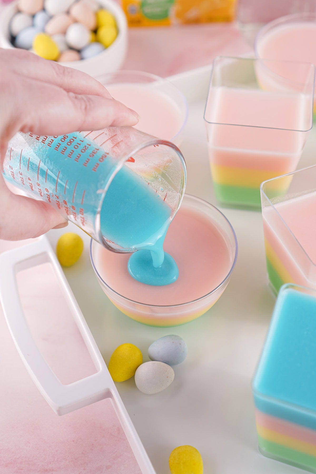 A hand carefully pouring a creamy blue jello layer from a measuring cup over a set pink layer in a round dessert bowl.