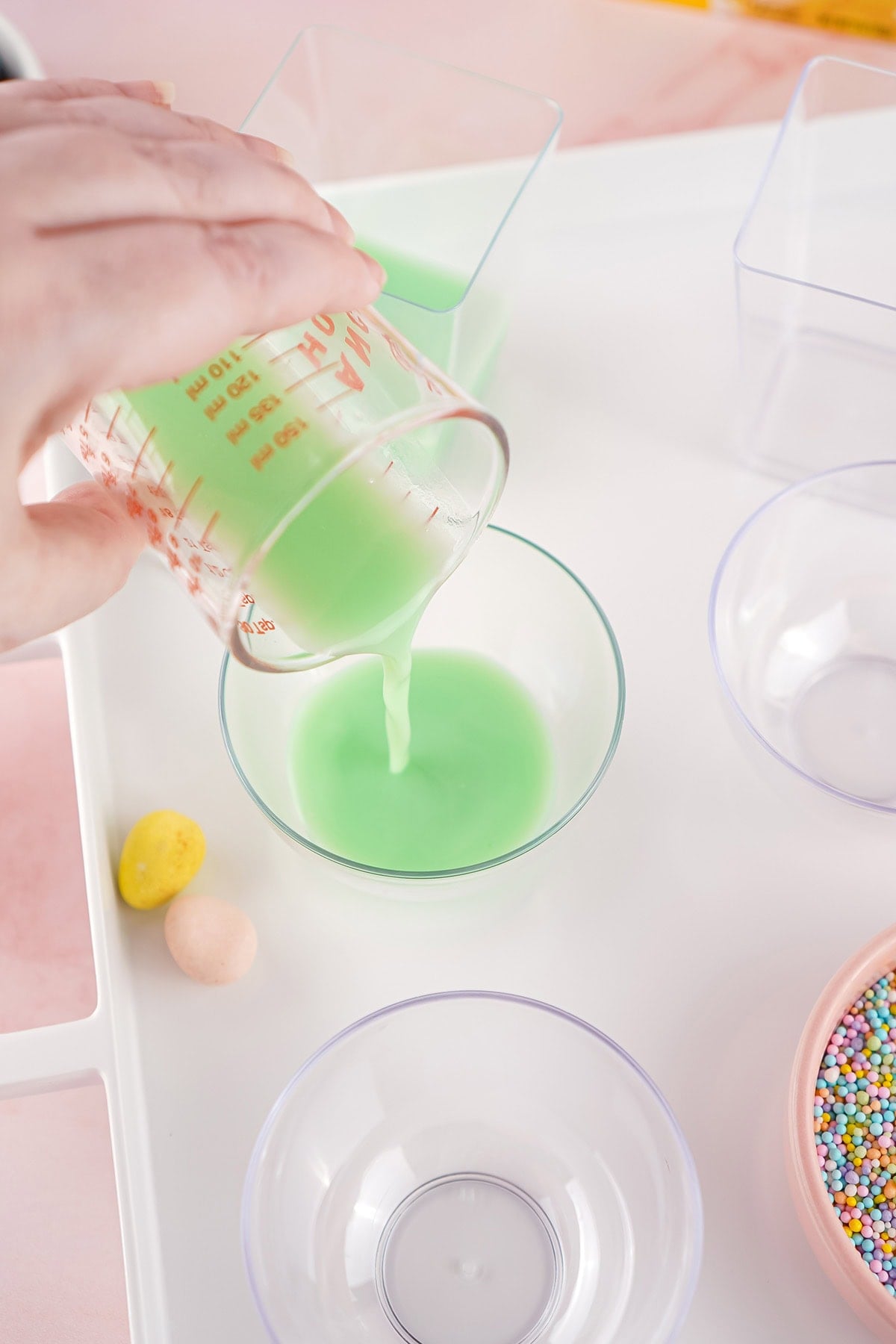 A hand pouring creamy green jello from a glass measuring cup into a small round dessert bowl to create the first pastel layer.